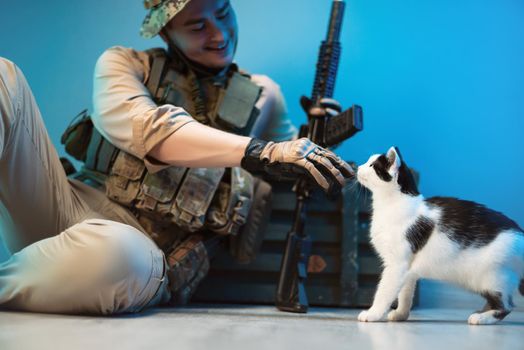 the a male soldier in camouflage is sitting on the floor by a box of ammunition with weapons and stroking a cat