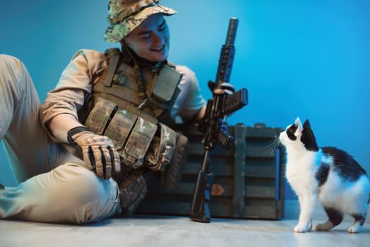 male soldier in camouflage is sitting on the floor by a box of ammunition with weapons next to a small cat