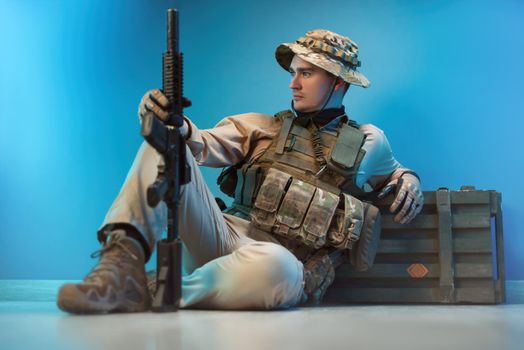 male soldier in camouflage is sitting by an ammunition crate on the floor with a weapon