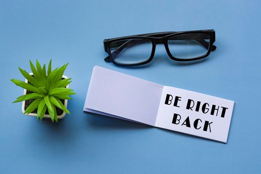 Be right back text on notepad with reading glasses and potted plant. Directly above. Flat lay, Message and reminder concept.