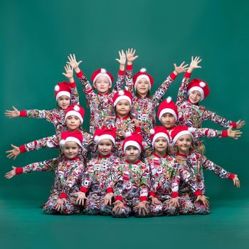 A group of children pose with outstretched arms. A team of dancers boys and girls in colorful Christmas costumes and Santa Claus hats pose together on a green background