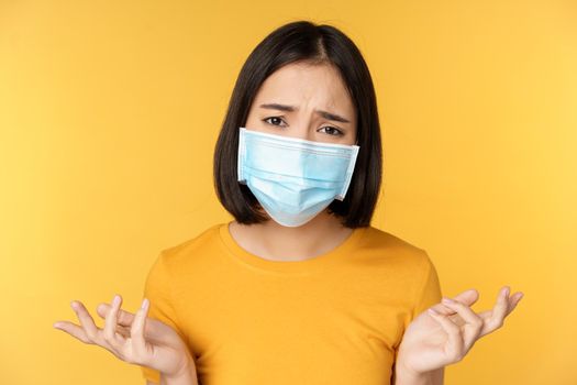 Close up portrait of confused asian woman in medical face mask, shrugging shoulders and looking puzzled, standing against yellow background.