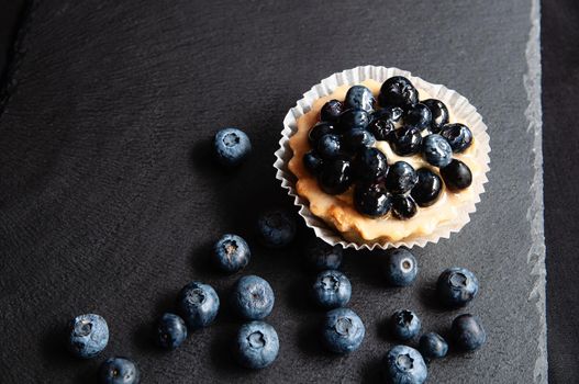 Tart with blueberries is on a black background of natural stone. Still life with sweet cakes with wild berries on a dark background