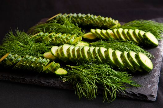 Natural fresh green cucumbers from a home garden on a black background, a dummy board made of stone. The cucumbers are cut into pieces and arranged in a dill pattern.