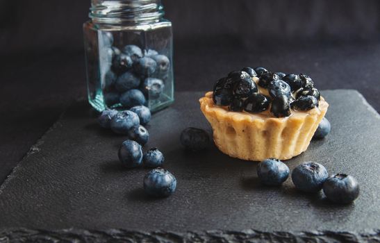 Blueberry tart on a dark background. Blueberry cupcake on black serving board with scattered berries