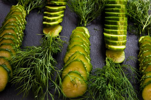 Natural fresh green cucumbers from a home garden on a black background, a dummy board made of stone. The cucumbers are cut into pieces and arranged in a dill pattern.