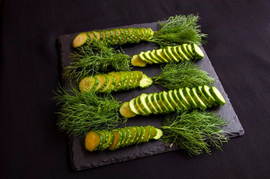 Natural fresh green cucumbers from a home garden on a black background, a dummy board made of stone. The cucumbers are cut into pieces and arranged in a dill pattern.