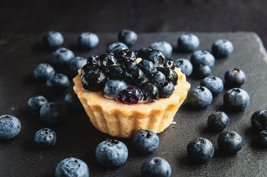 Tart with blueberries is on a black background of natural stone. Still life with sweet cakes with wild berries on a dark background