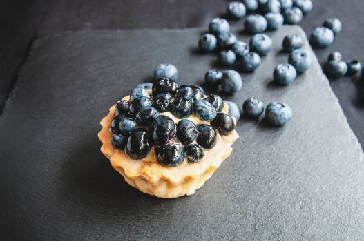 Delicious and sweet tarts made from cream and blueberry. Blueberry tart on a dark background. Blueberry cupcake on black serving board with scattered berries