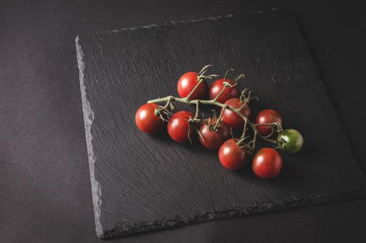 bunch of fresh cherry tomatoes on a branch are located on a black serving board on a dark background