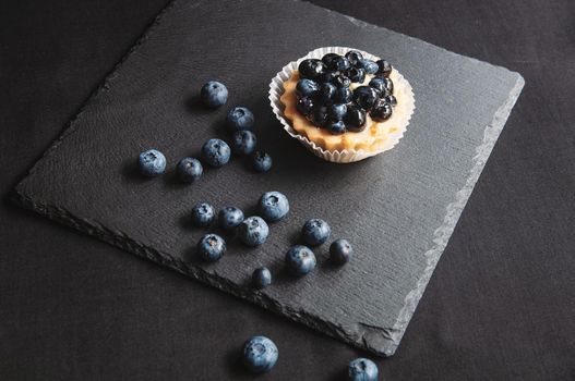 Tart with blueberries is on a black background of natural stone. Still life with sweet cakes with wild berries on a dark background