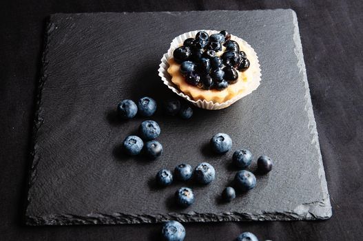 Tart with blueberries is on a black background of natural stone. Still life with sweet cakes with wild berries on a dark background