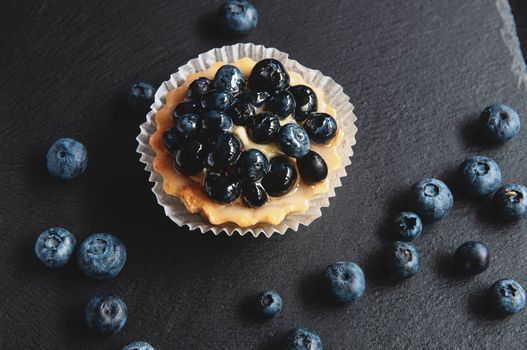 Tart with blueberries is on a black background of natural stone. Still life with sweet cakes with wild berries on a dark background