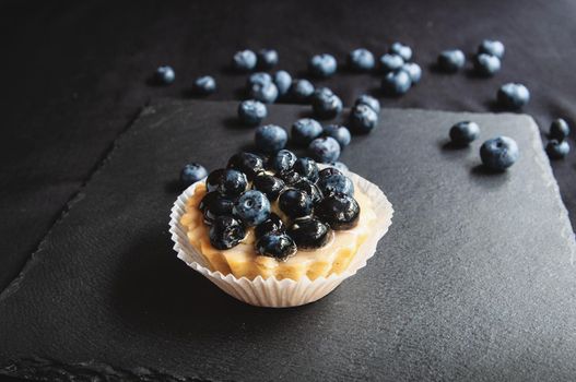 Delicious and sweet tarts made from cream and blueberry. Blueberry tart on a dark background. Blueberry cupcake on black serving board with scattered berries