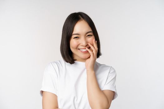 Portrait of young beautiful woman, korean girl laughing and smiling, looking coquettish, standing against white background.