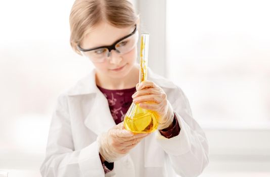 Smart girl during scientific chemistry experiment wearing protection glasses, holding bottle with orange liquid. Schoolgirl with chemical equipment on school lesson portrait