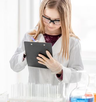 Smart girl during scientific chemistry experiment wearing protection glasses making notes. Schoolgirl with chemical equipment on school lesson