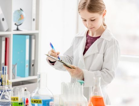 Girl during scientific chemistry experiment making notes close to table with tubes. Schoolgirl with chemical equipment on school lesson