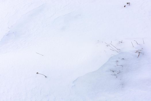 Snowy uneven white background with dry grass showing in places. Winter meadow. View from above.