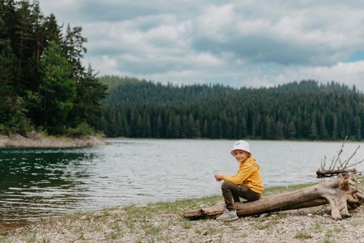 Little boy sitting on the log near the mountain Lake. A lonely boy sits quietly and looks ahead at a beautiful view of mountains. little boy lake mn, little boy lake resort.