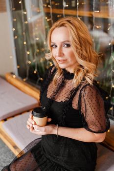 Young charming blonde with a cute smile and makeup while relaxing in a cafe. She is holding a cup of coffee in her hands. She is dressed in a black dress with transparent sleeves