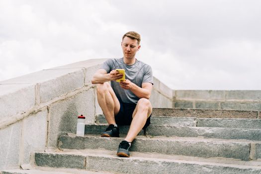 Tired Young man runner sitting on stairs, using mobile phone and relaxing after sport training. Holding water bottle while doing fitness workout in city urban street, cloudy sky at summer.