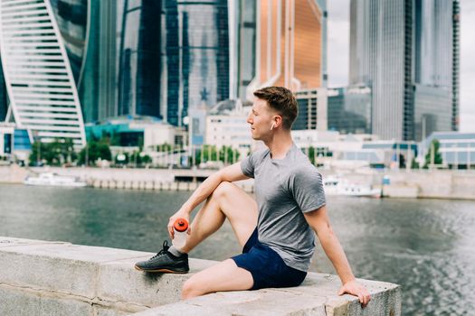 Tired Young man runner sitting on stairs and relaxing after sport training. Holding water bottle while doing fitness workout in summer city urban street, cloudy sky