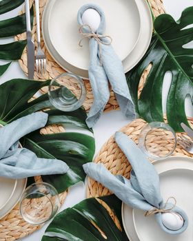 Tropical Easter Tablesetting. Top view of table setting for Christian Easter with bunny eggs decorated monstera leaves. Aesthetic Easter tablescapes. Easter dinner with bunny, tropical leaves and eggs