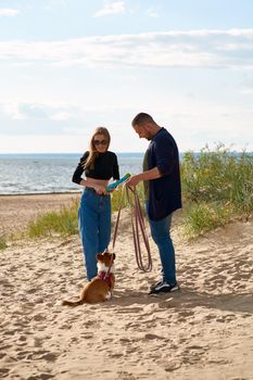 Young happy couple playing with dog by toy. People walking along beach with pet. Man holding Corgi puppy on leash. Summer walk in nature of family in love on weekend. Full length peoples