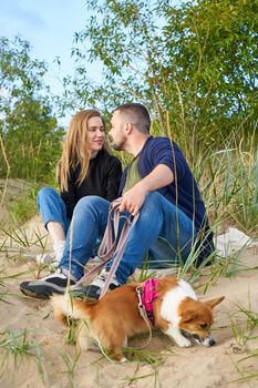 Young happy couple of man and woman with corgi dog sit at sand. Male kissing beautiful female. Two persons in casual clothes enjoing beautiful sunny afternoon on beach