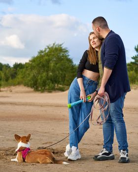 Young happy couple and dog stand on beach against pines and sand. Handsome man gently kissing woman while holding Corgi puppy on leash. Walking in summer nature of people in love