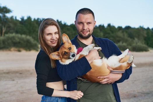 Young happy couple with dog standing on beach. Beautiful girl and guy and Corgi puppy having fun. Family life, togetherness, husband and wife relax together in summer by sea