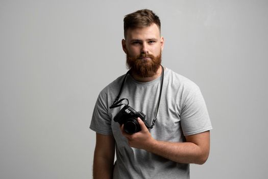 Portrait of bearded professional photographer with dslr camera looks straight into the camera isolated on gray background