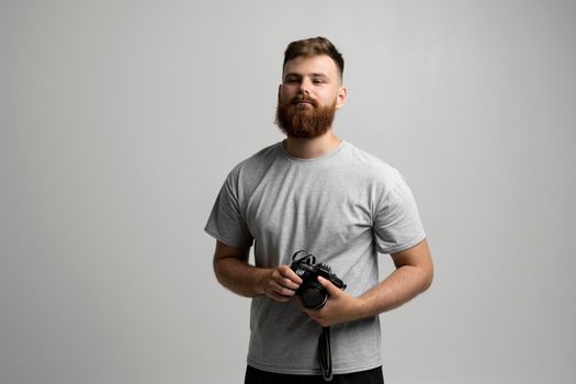 Portrait of bearded professional photographer in a grey t-shirt with dslr camera looks straight into the camera isolated on gray background