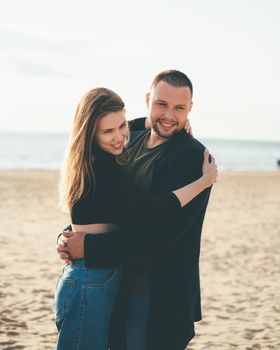 Young adult couple standing on coast and huging each other and looking away to someone. Handsome smiling man embracing beautiful smiling woman. Millennial male and female posing on beach