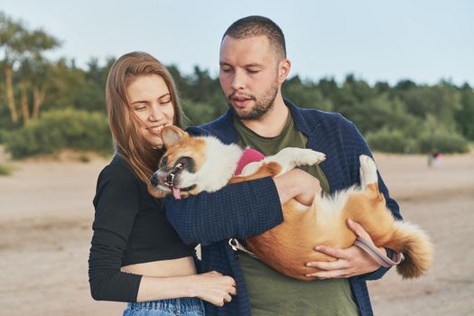 Young happy couple with dog standing on beach. Beautiful girl and guy and Corgi puppy having fun. Family life, togetherness, husband and wife relax together in summer by sea