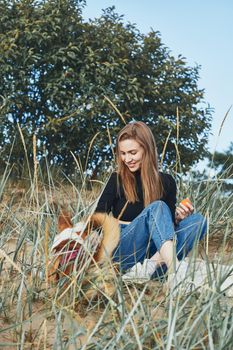 Full length beautiful pensive woman and dog sitting on beach at sunset in evening. Pretty female with long brown hair, looking to pet corgi. Slow life, social detox