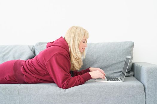 Mature woman working at home with laptop laying on comfortable sofa, room interior. Beautiful female shopping or chatting online in social network, freelancer working