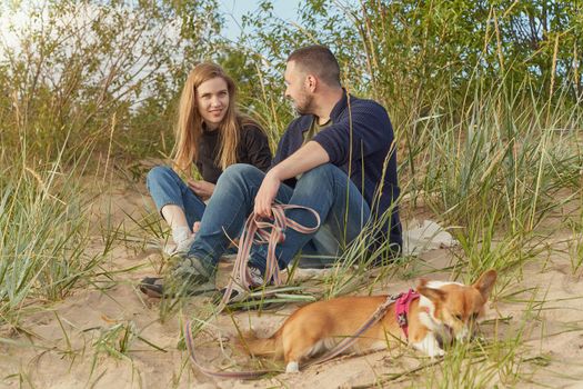 Young happy couple with corgi dog, man and woman siting at sand. Two persons in warm casual clothes talking in beautiful sunny afternoon on beach
