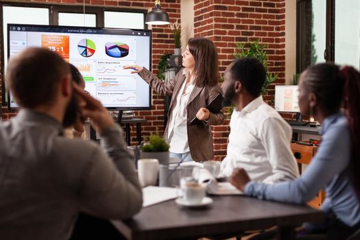 Entrepreneur woman standing in front on monitor explaining marketing strategy working at business presentation in startup company office. Diverse businessteam brainstorming project ideas