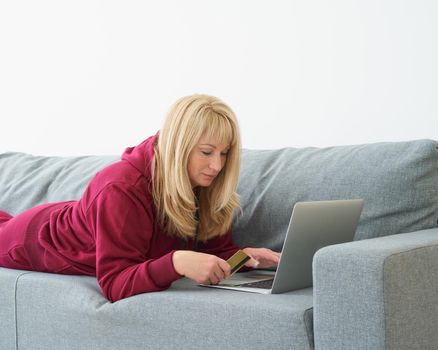 Mature woman online shopping, lying on couch with laptop and credit card. Middle-aged female in bright red sportswear paying for purchases