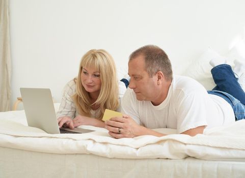 Mature family couple sitting at bed in modern home, looking at laptop screen. Happy middle aged man holding credit card and female making purchases online