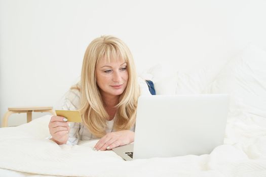 Middle age female on online shopping. Portrait of Mature beautiful blond woman is holding credit card and using laptop computer laying on bed at home