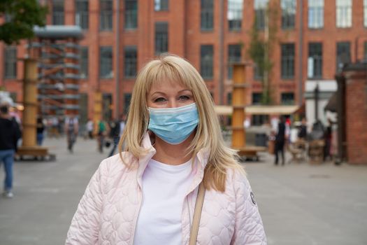 Woman in protective medical face mask standing outside in public place outdoor, looking at camera. Mature middle-aged female