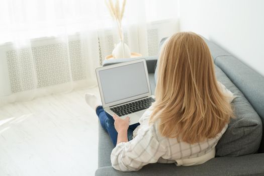 Back view of unrecognizable mature woman using laptop while sitting on comfortable sofa, home interior. Beautiful female shopping or chatting online in social network, freelancer working