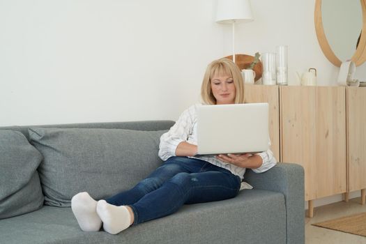 Mature woman using laptop while sitting on comfortable sofa, home interior. Full length beautiful female shopping or chatting online in social network, watching movie, freelancer working