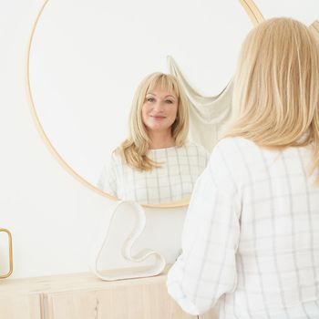 Middle-aged woman looks at herself. Mature beautiful blonde woman with long hair admires reflection standing in front of large round mirror in apartment of home, looking to camera