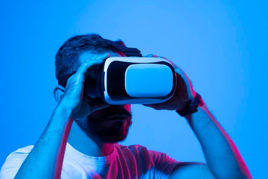 Closeup of young man in white t-shirt wearing virtual reality goggles in modern studio with a neon light