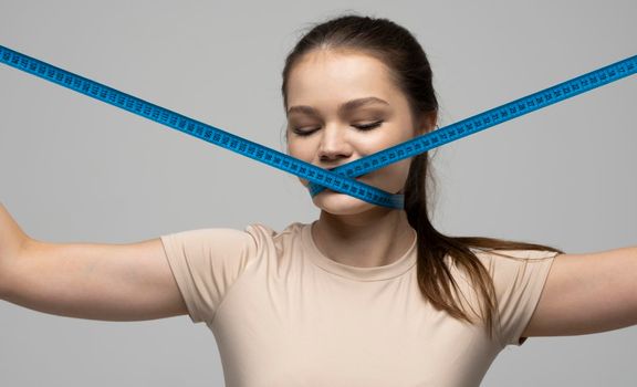 Young attractive beautiful brunette woman in a beige outfit holding measuring tape around her mouth on white background. Healthy lifestyle and diet concept