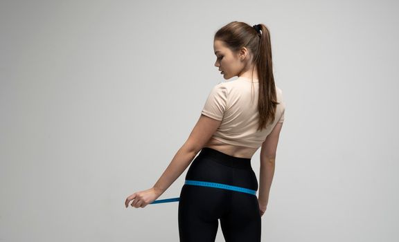 Young brunette woman in sportswear holding measuring tape around her thighs on white background. Weight loss and diet concepts